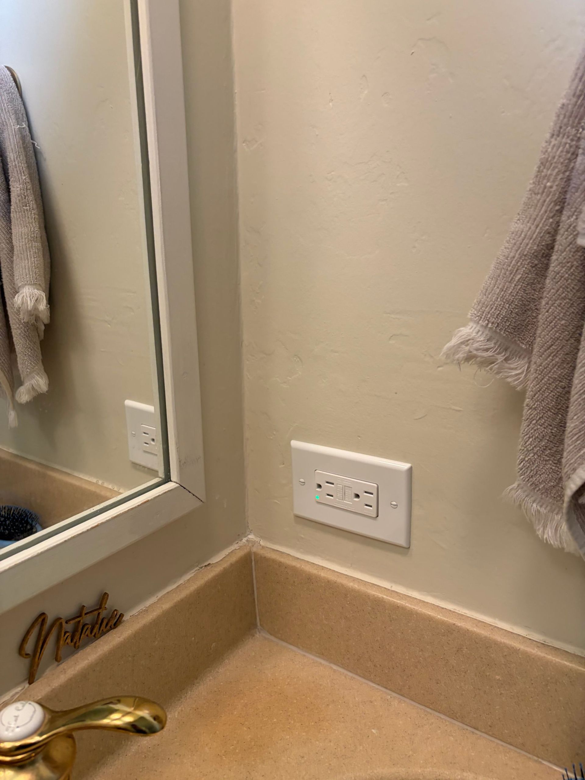 Bathroom corner with mirror, gold faucet, beige walls, towel racks, and a wall outlet.