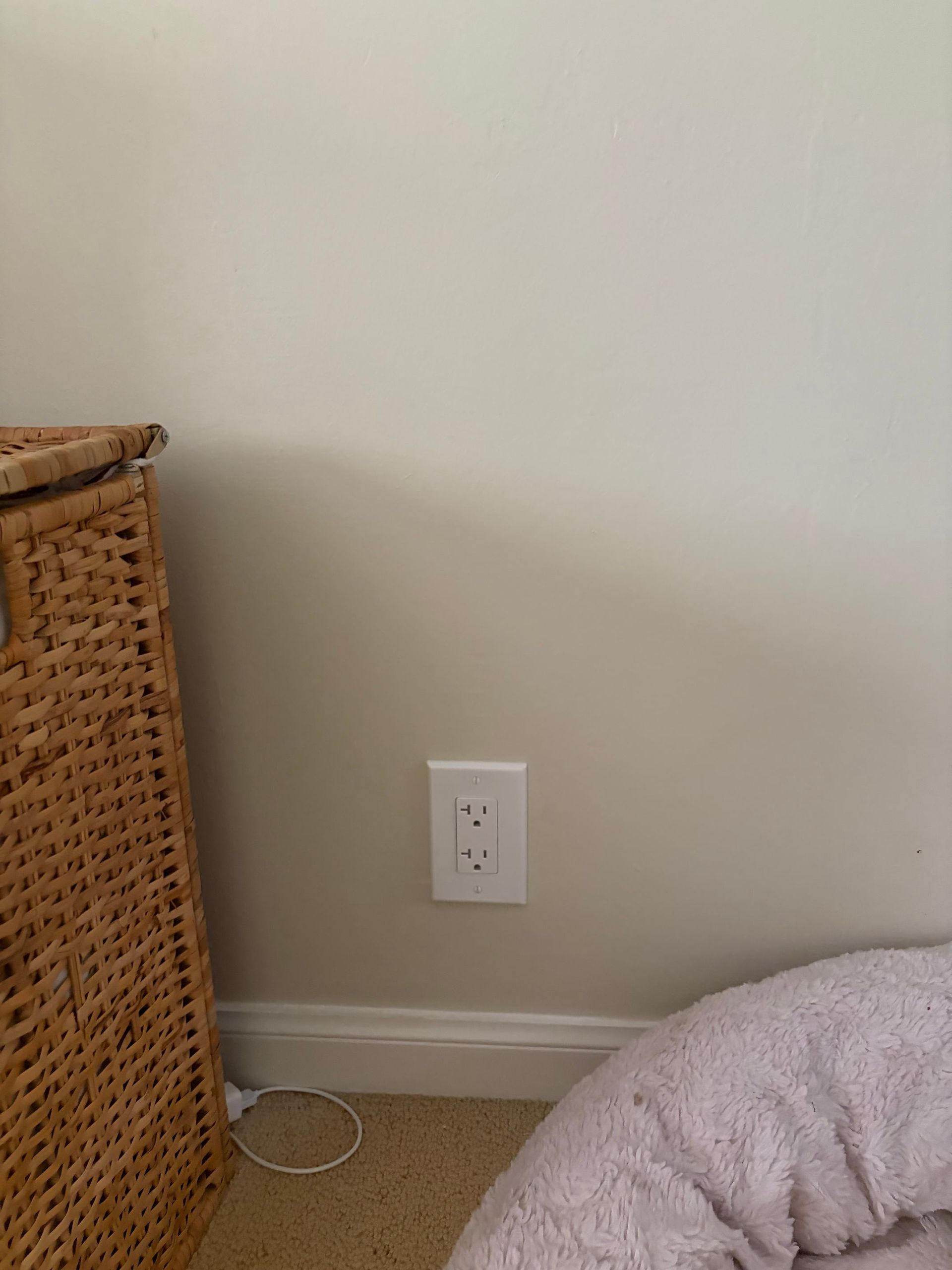 Wall outlet on a beige wall beside a wicker basket and carpeted floor.