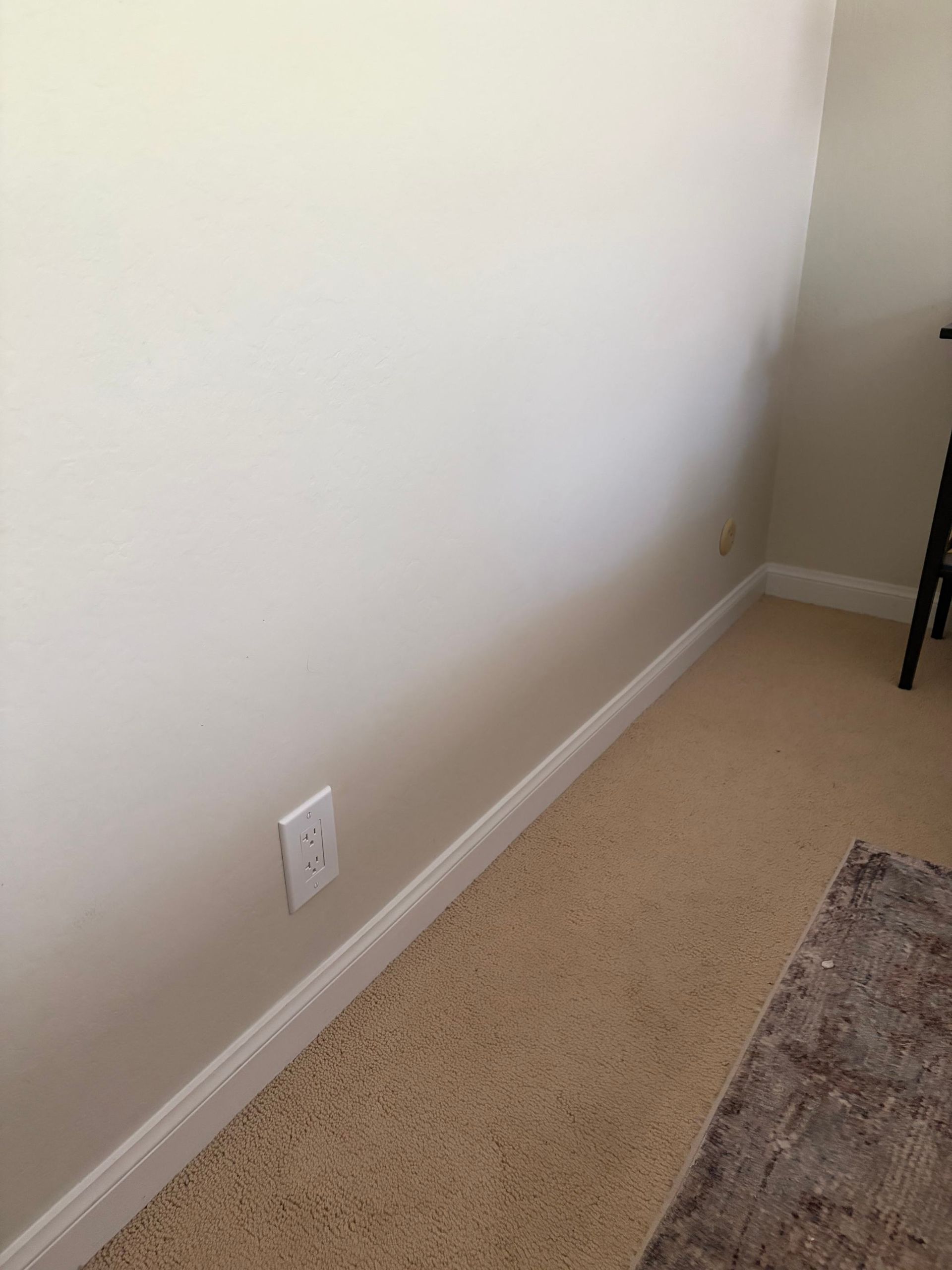Empty beige room corner with white baseboard, outlet, and partial rug on carpeted floor.