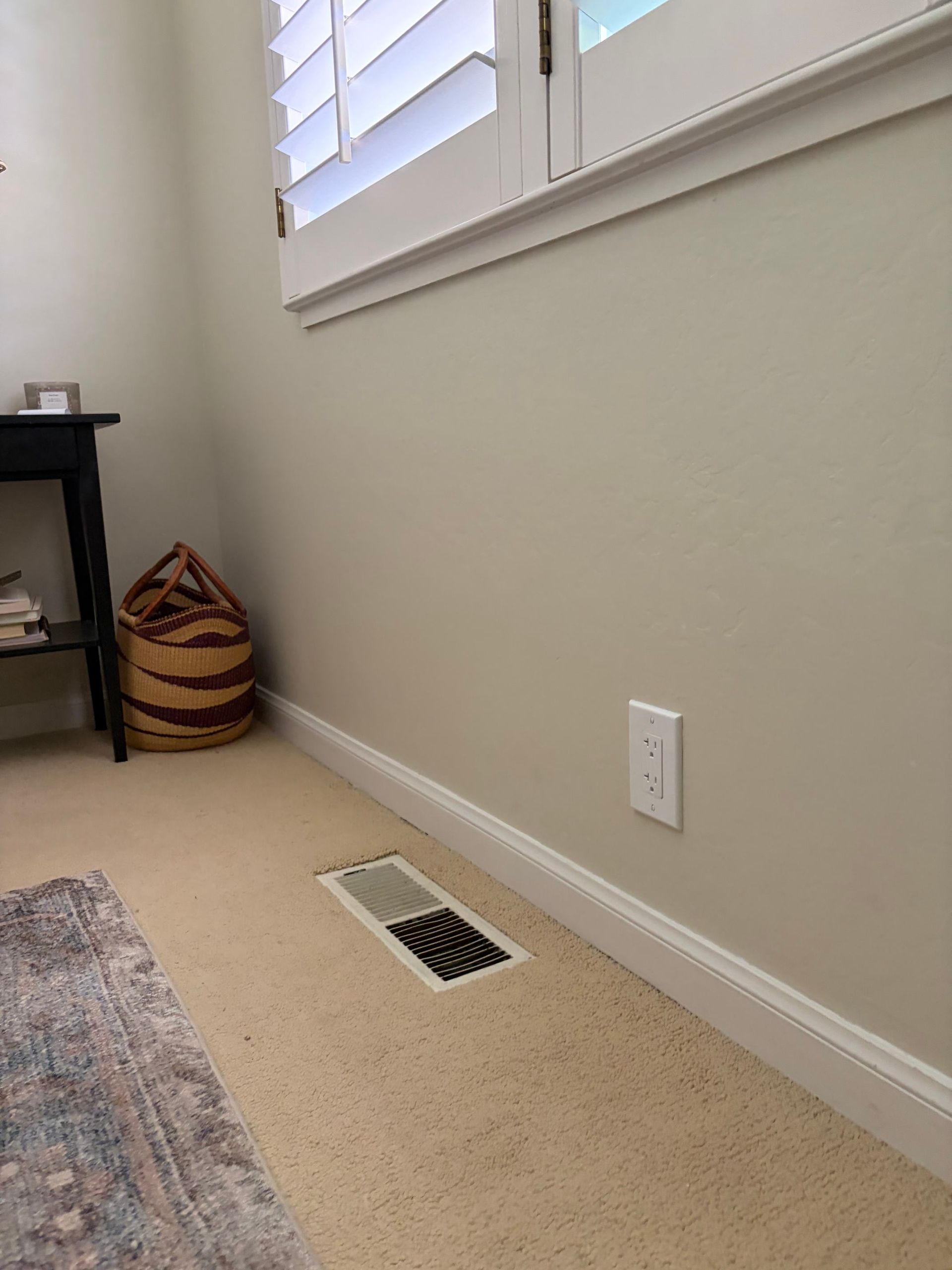 Bedroom corner with beige carpet, wall vent, white outlet, window, and a woven basket by a side table