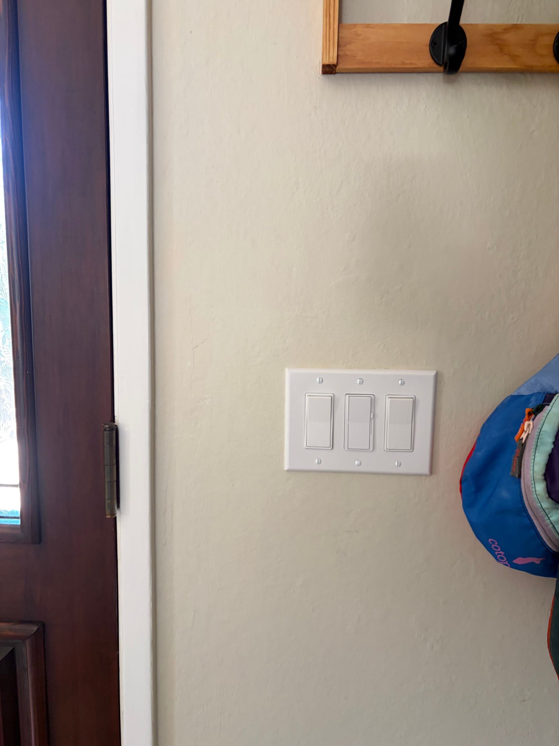 White wall with a light switch panel beside a dark wooden door and a blue hat on the right.