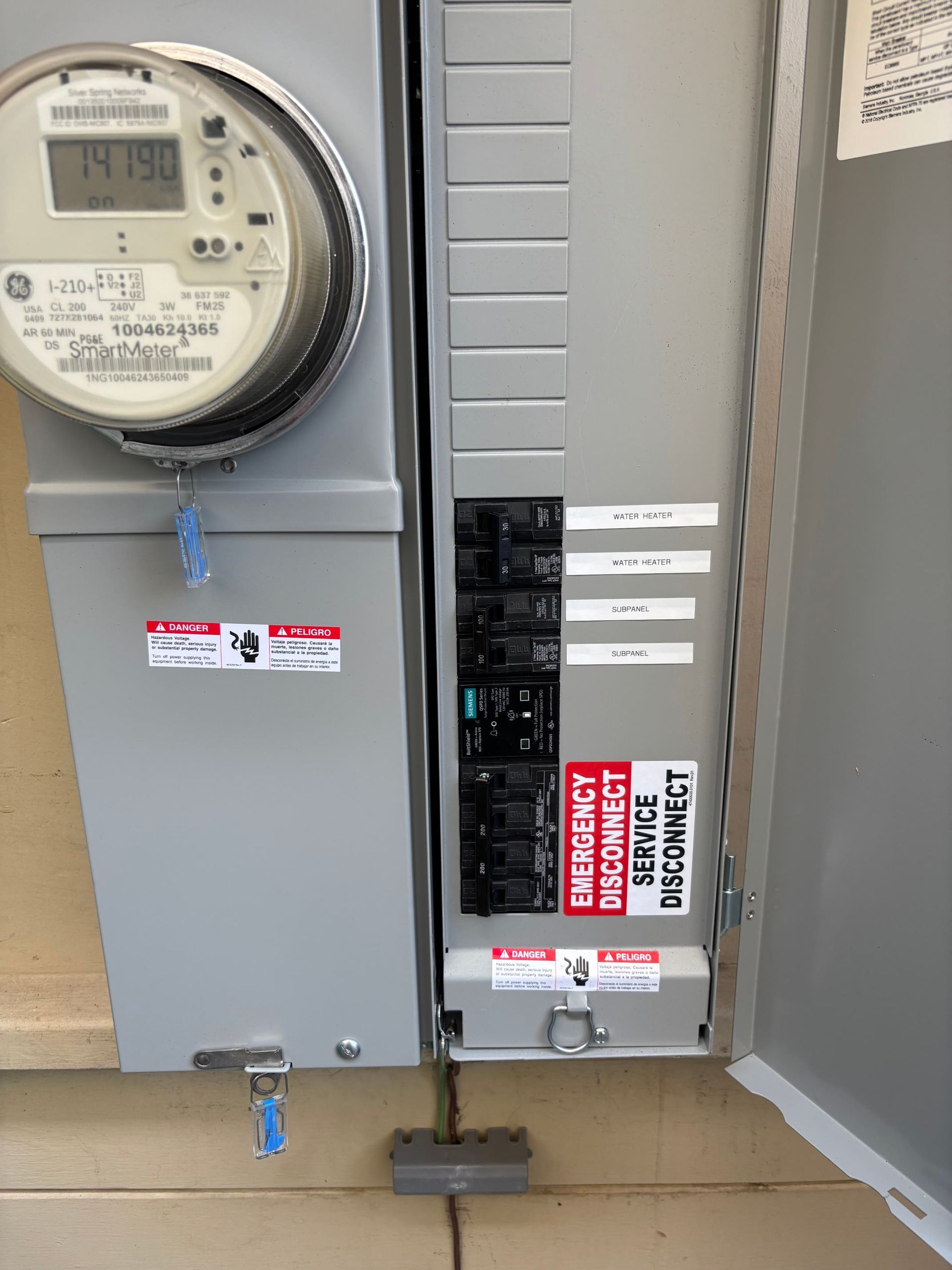 Electric meter and open breaker panel on a gray wall, with labels and warning stickers.
