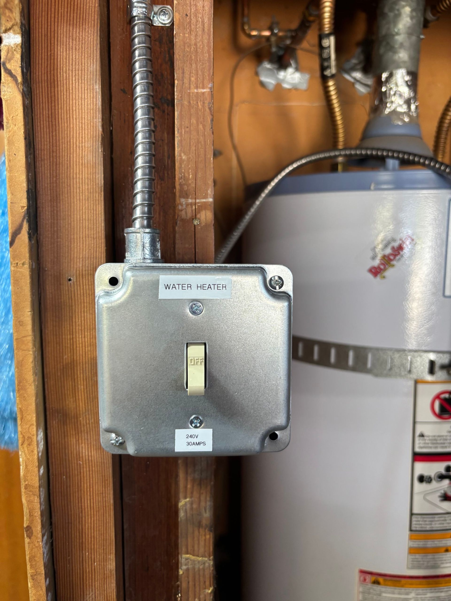 Silver switch box mounted on a wooden wall beside a water heater and exposed pipes.