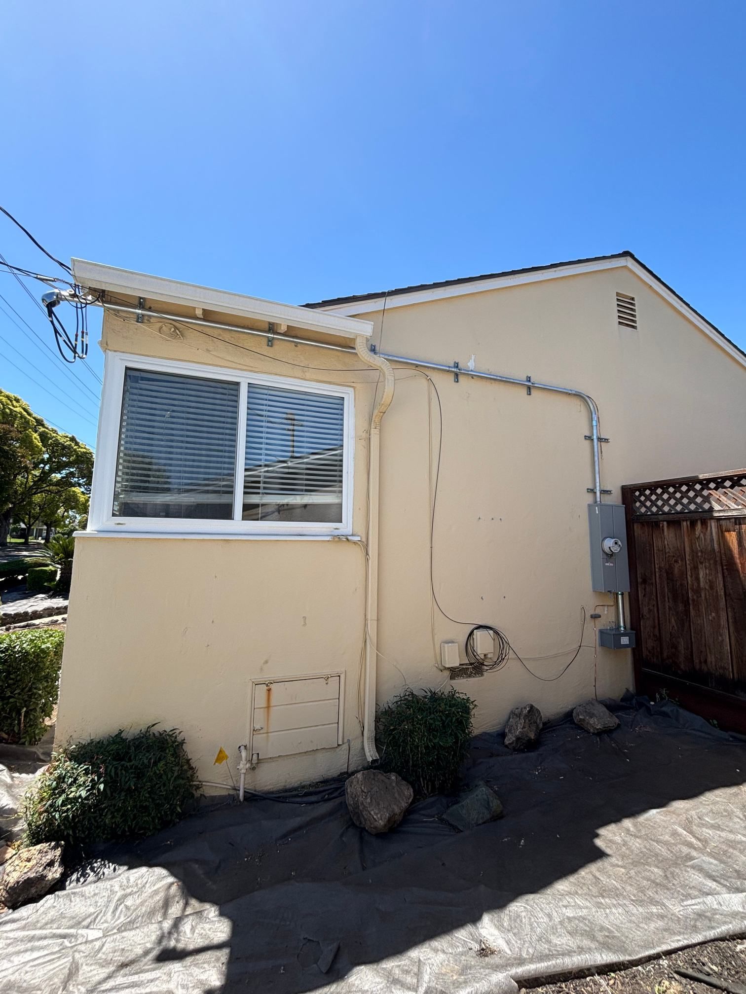 Side of a beige house with a window, utility pipes, and a stone walkway under a blue sky
