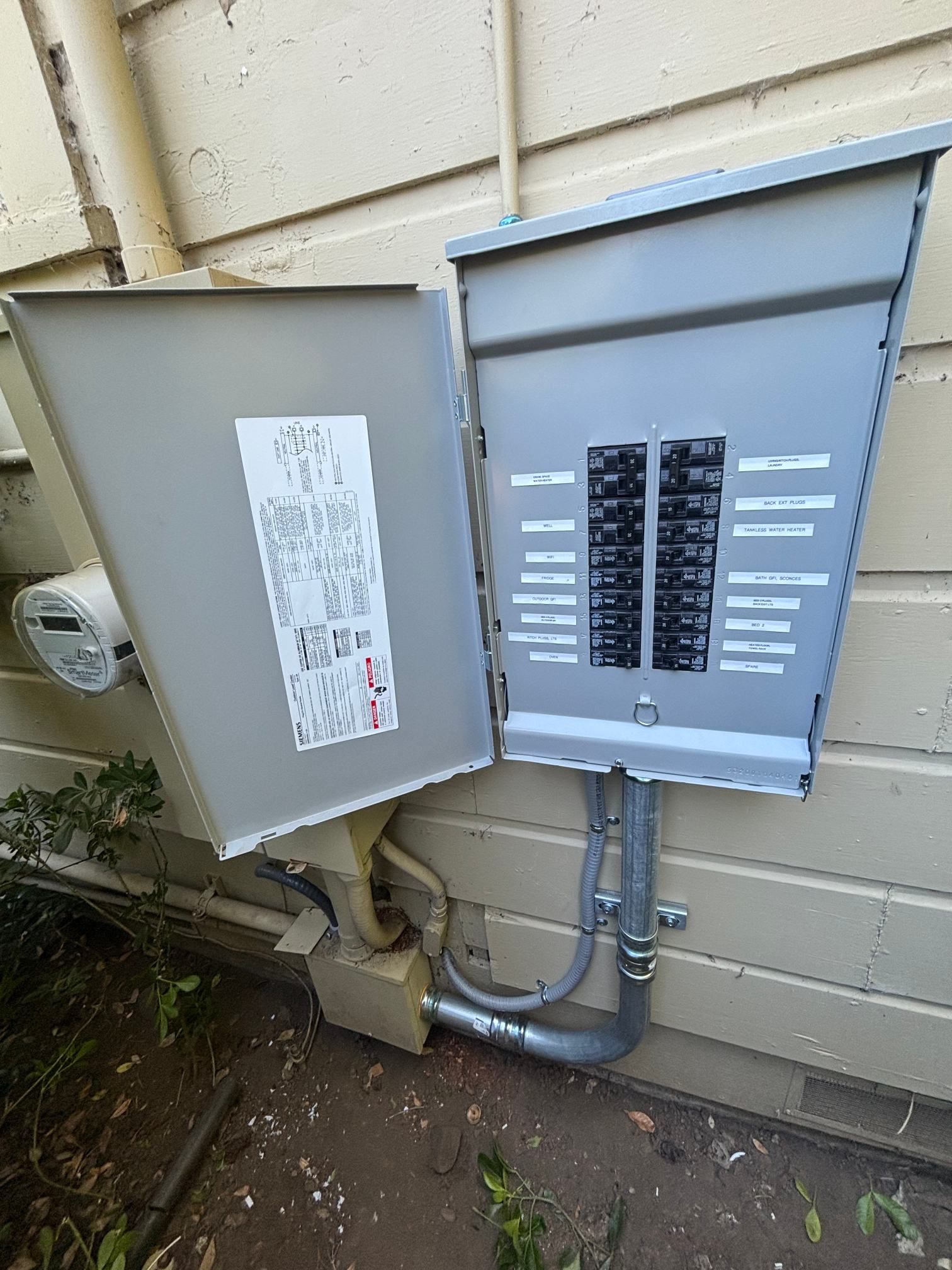 Open electrical breaker panels mounted on beige exterior siding with visible conduit below.