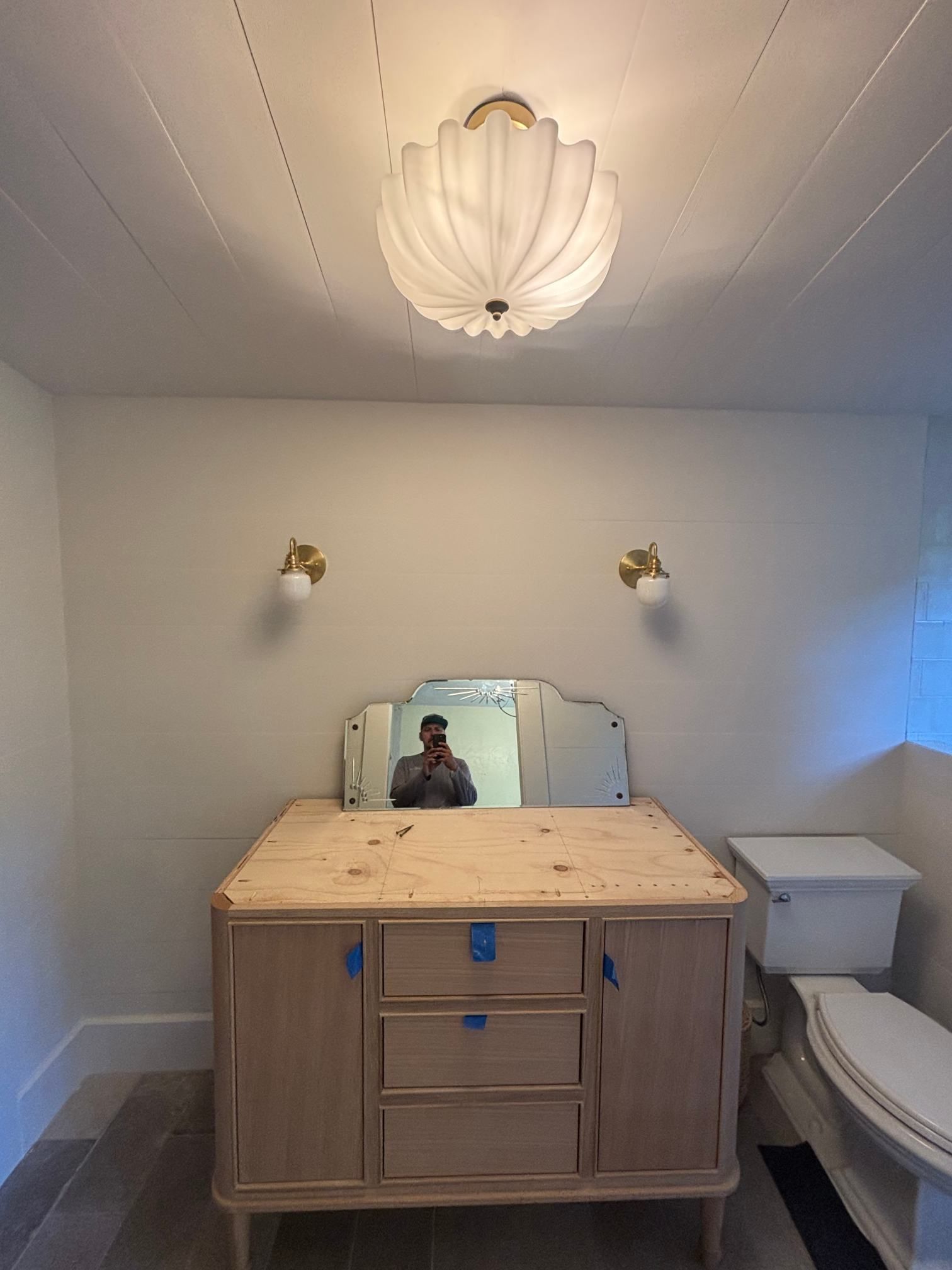 Small bathroom with a light wood vanity, mirror, twin wall lights, and a white toilet in the corner.