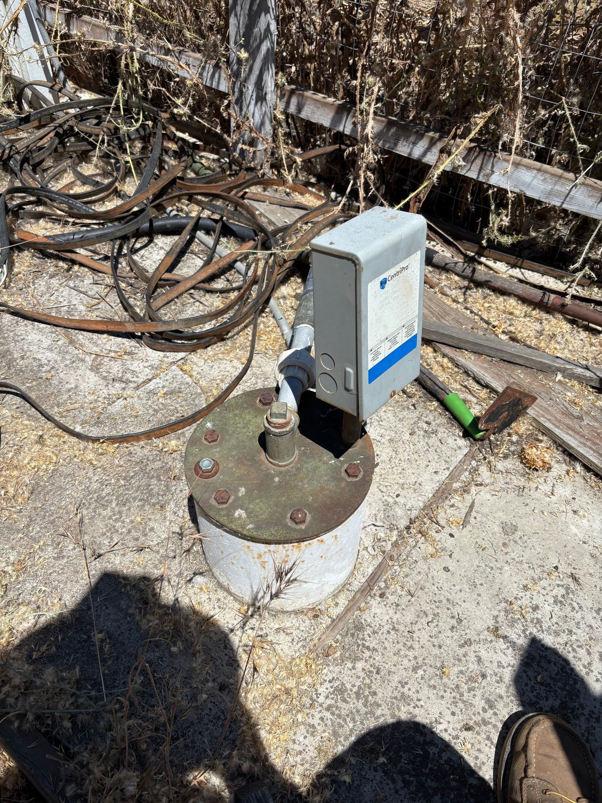 Electrical box mounted on a rusted metal plate outdoors, with tangled cables and dry brush nearby
