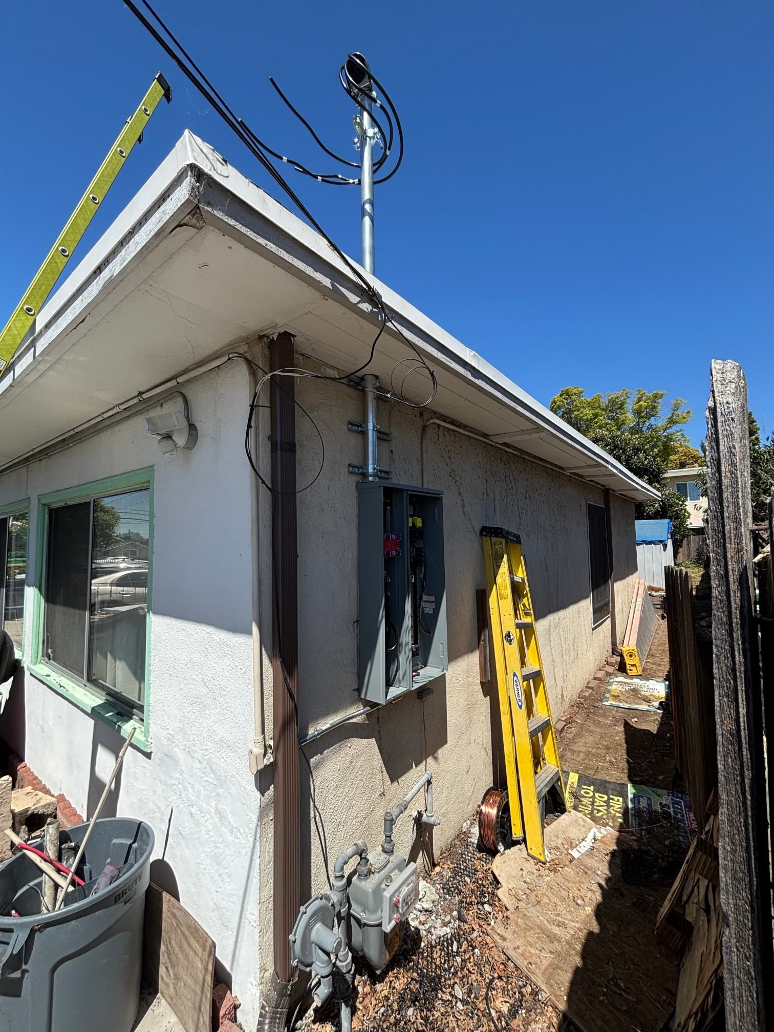 Narrow side yard beside a white house with utility poles, ladder, and construction materials under a blue sky