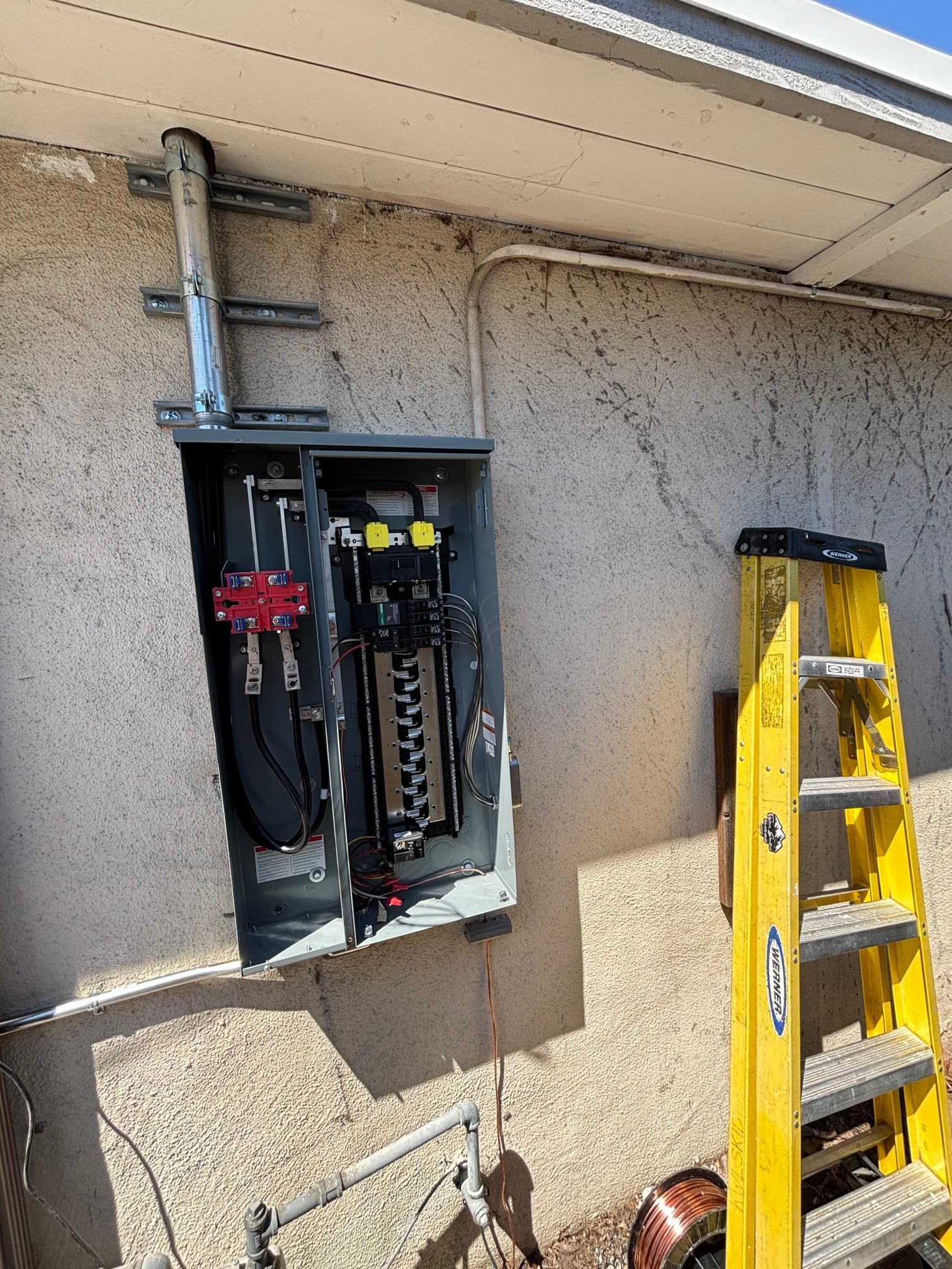 Open electrical panel on exterior wall beside a yellow ladder, with exposed wiring and conduit.