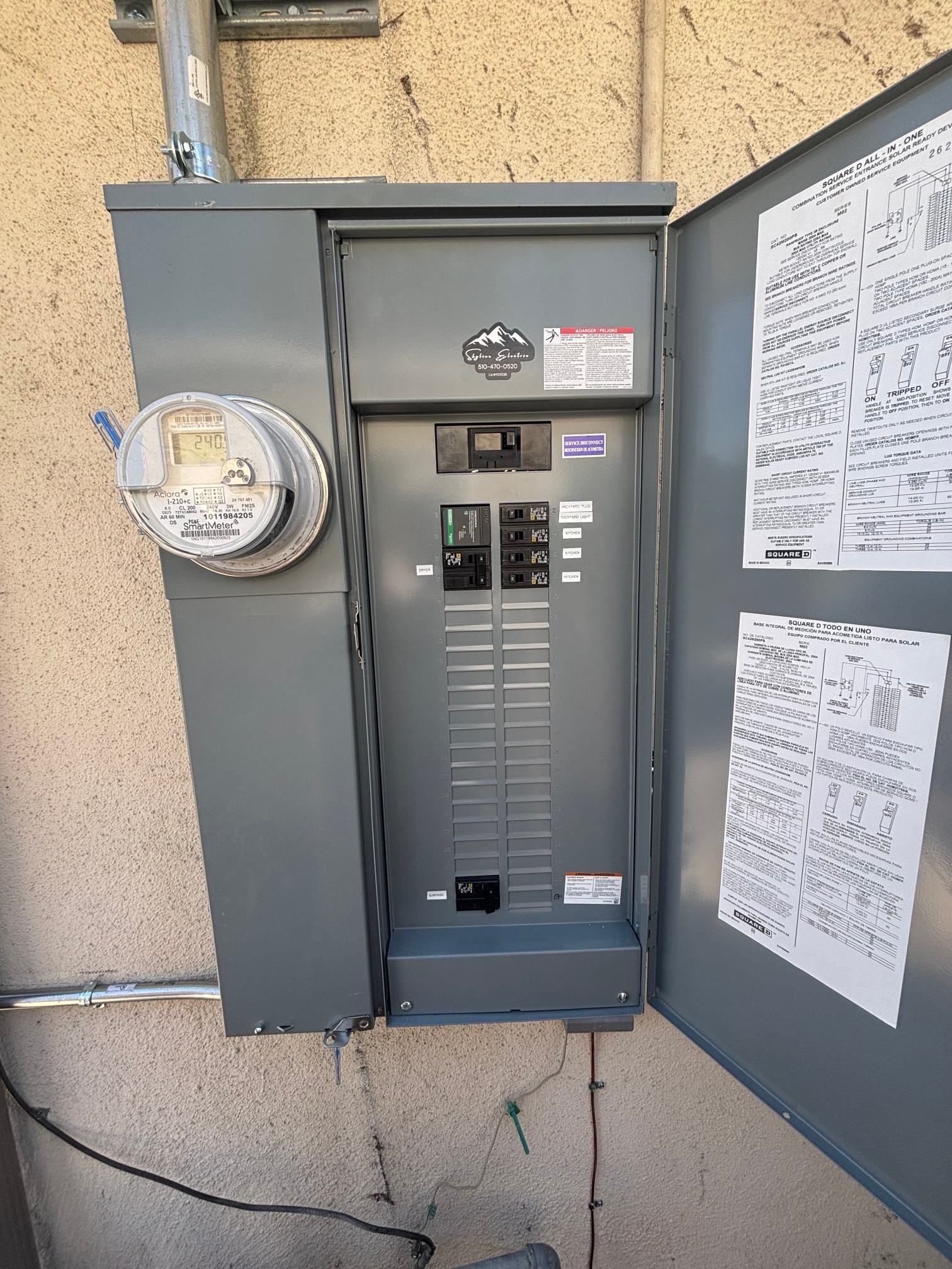 Open electrical breaker panel on exterior wall beside a utility meter and posted notices.