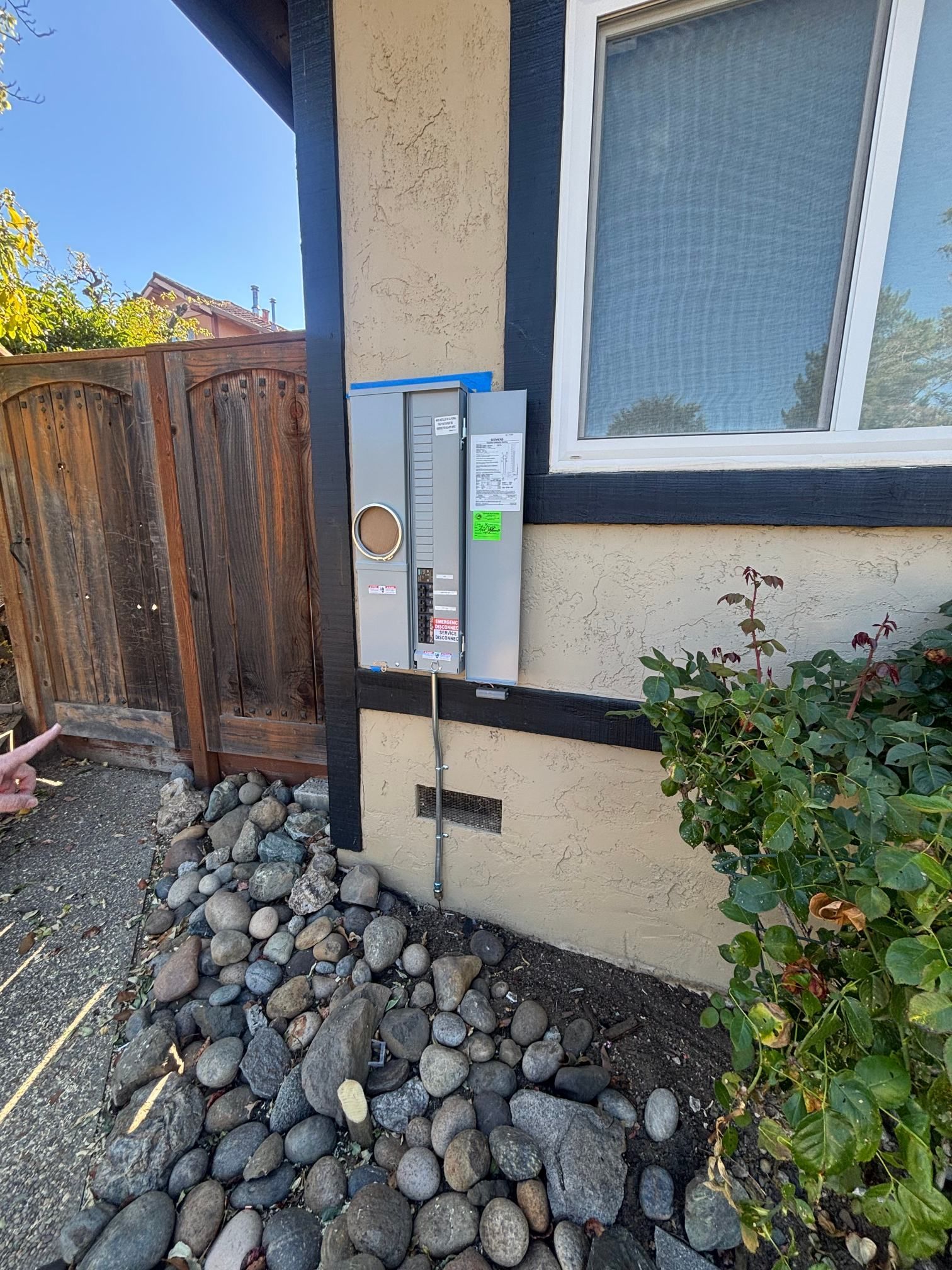 Outdoor electrical meter and service boxes mounted on a house wall beside a window and rock landscaping
