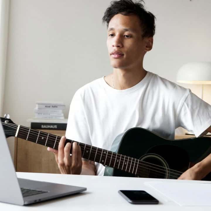 Man playing an acoustic guitar while using a laptop in a well-lit room, ideal for music creativity concepts.