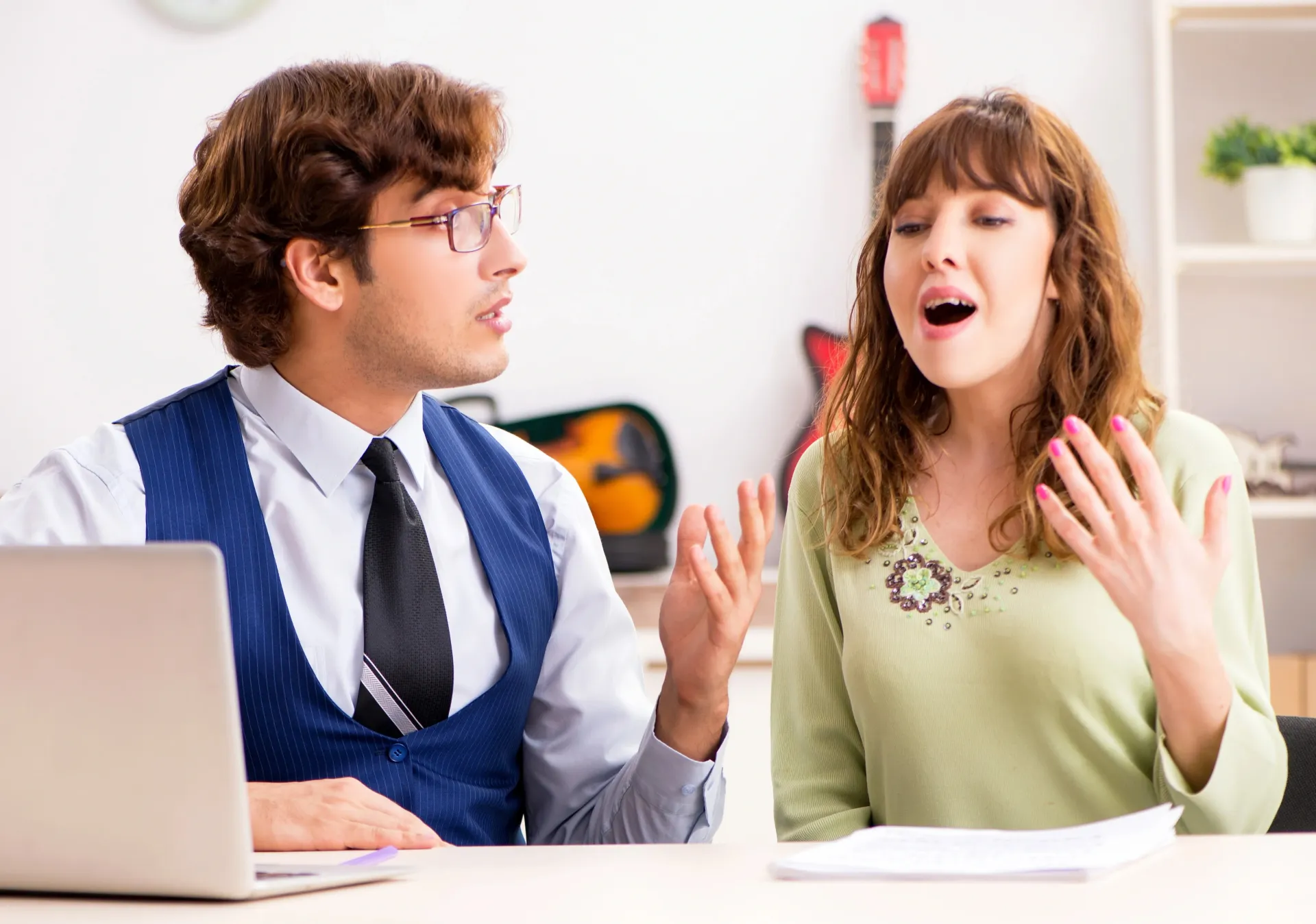 A music teacher is teaching a woman how to sing. They are in a well-lit office with a computer open.