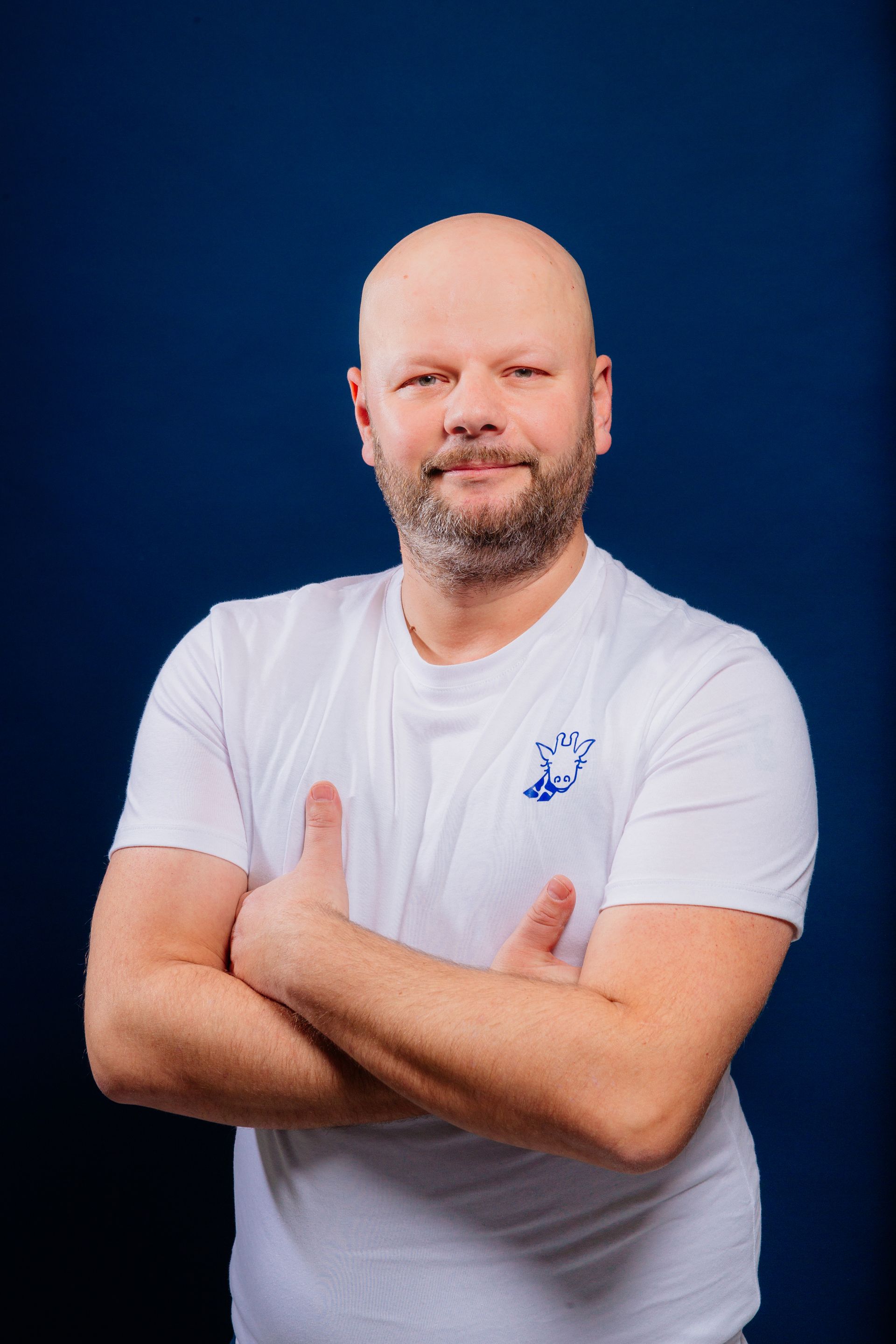 Un homme chauve avec une barbe porte une chemise blanche avec les bras croisés.