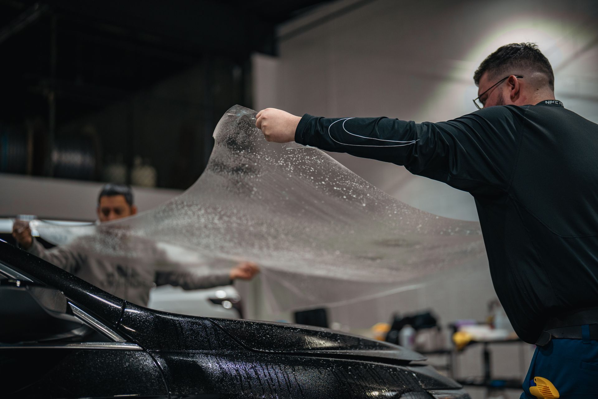 Two workers stretch a clear protective film over a black car in a garage