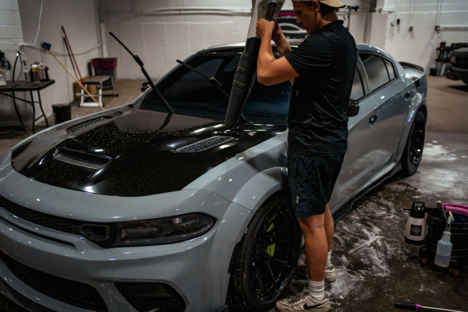 Person detailing a gray sports car in a garage, spraying the hood and windshield with water