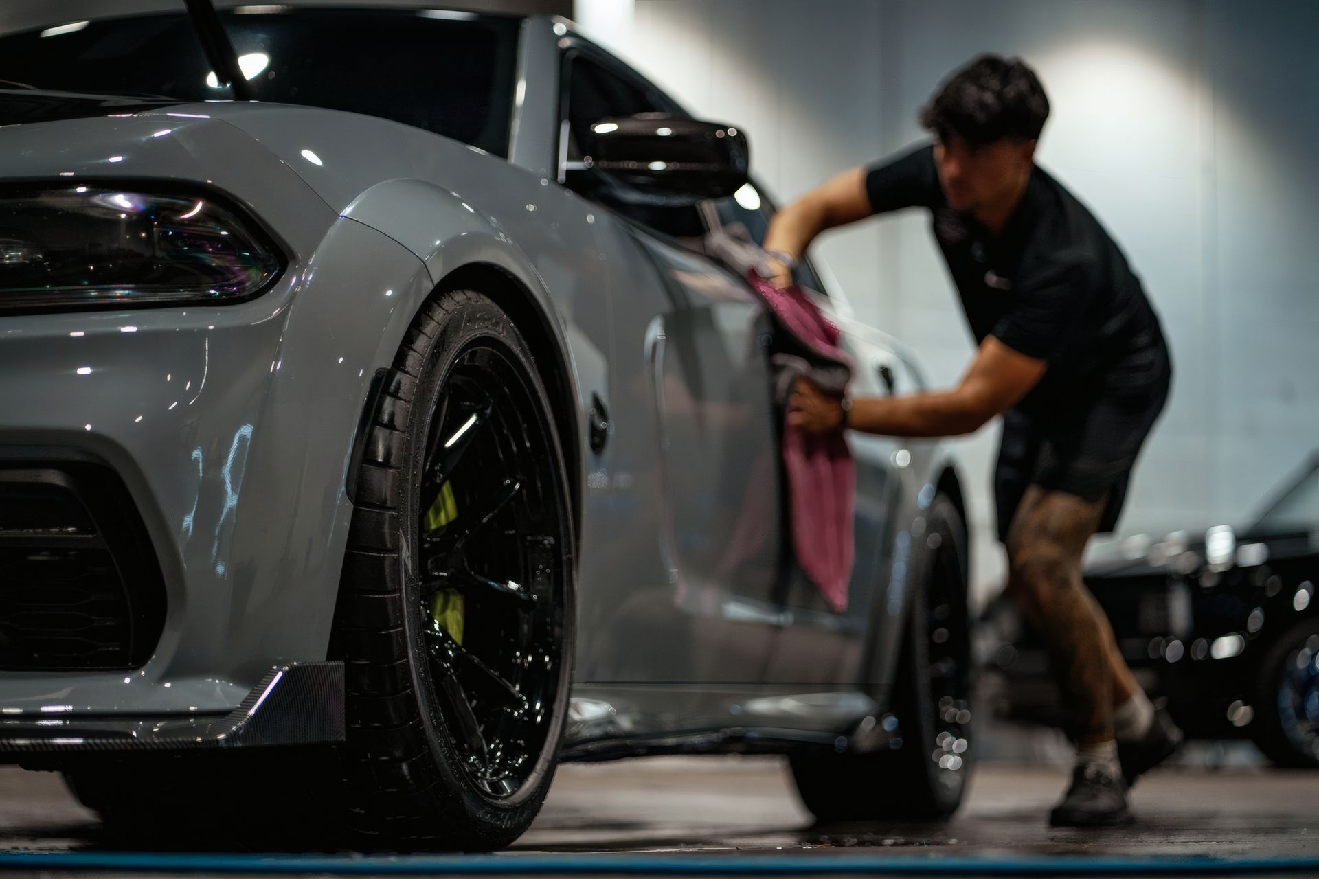 Person detailing a gray sports car in a garage, wiping the side with a cloth.