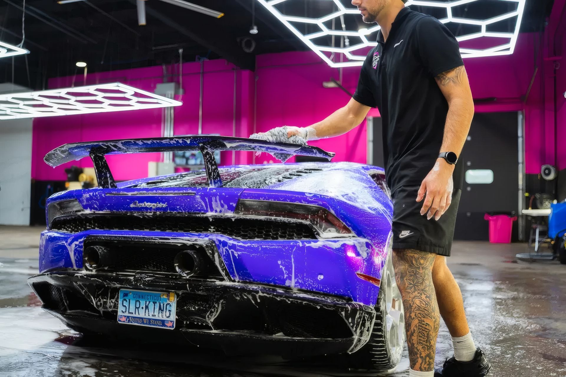 Person washing a blue sports car in a neon-lit car detailing shop