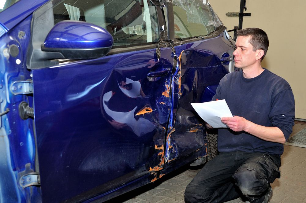A Man Inspecting a Damaged Blue Car Door With a Checklist in a Garage — Kingston Panel & Paint in Huntingfield, TAS