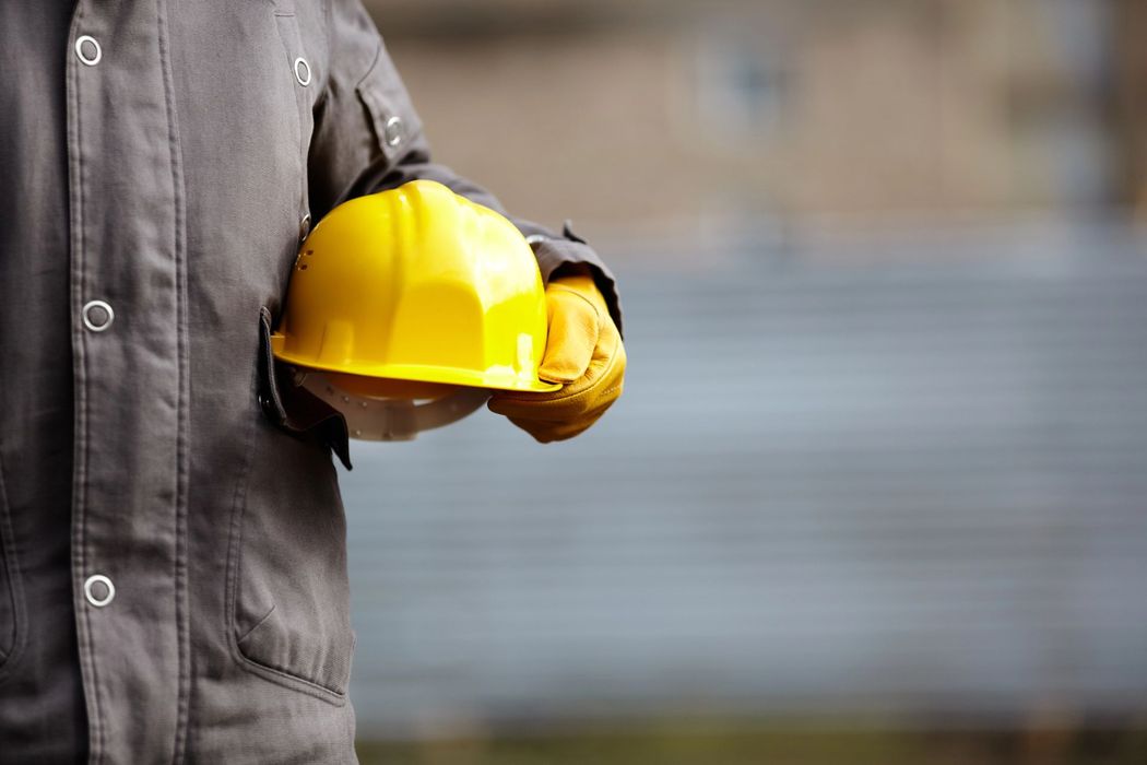 Persona con indosso un elmetto giallo e una giacca da lavoro grigia, sullo sfondo di uno sfondo industriale sfocato.