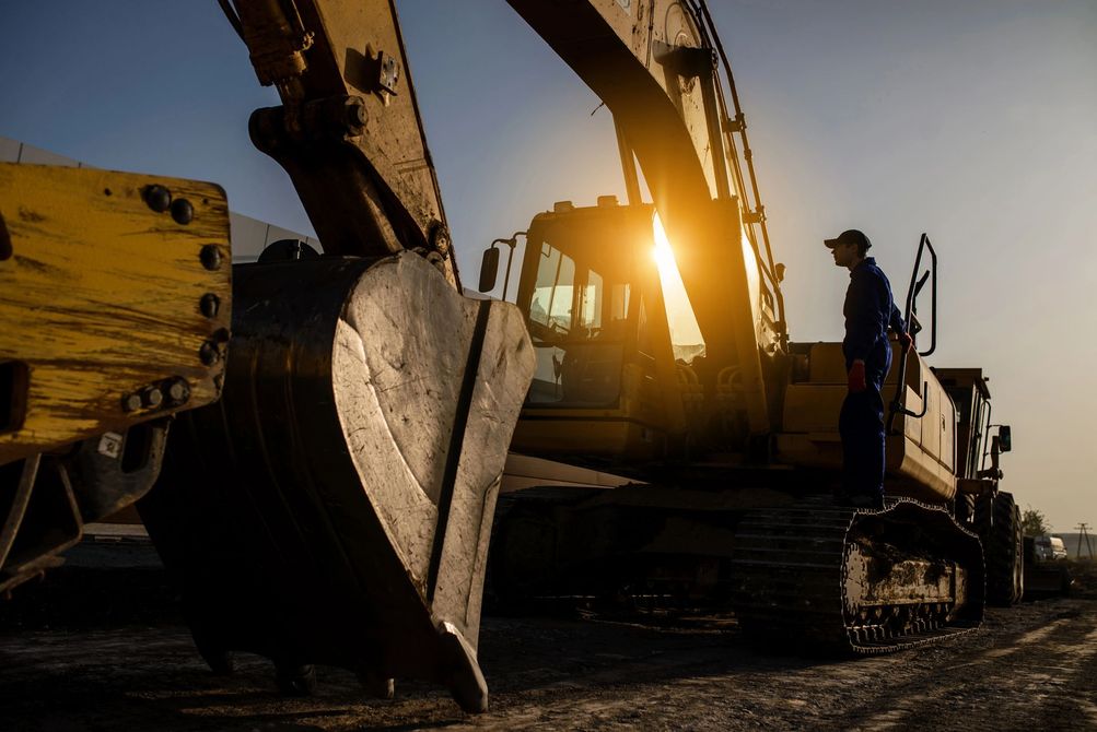 Un escavatore al tramonto in un cantiere, con un operaio in piedi accanto.