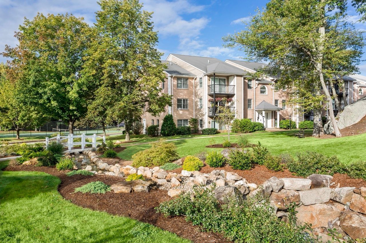 Exterior view of a brick apartment complex with landscaped lawn, trees, and stone garden beds.