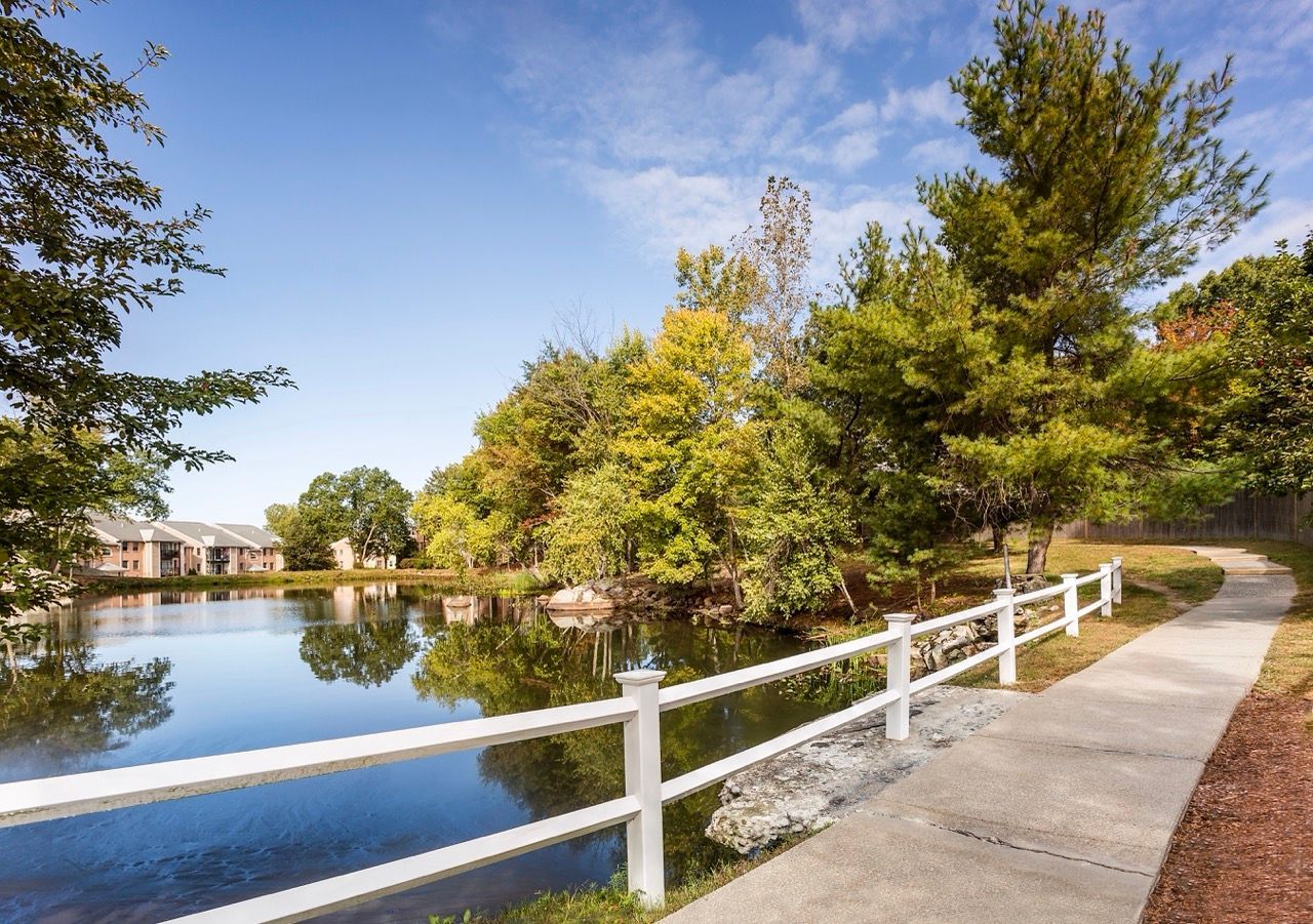 Path along a pond with trees and a white fence at a residential community.