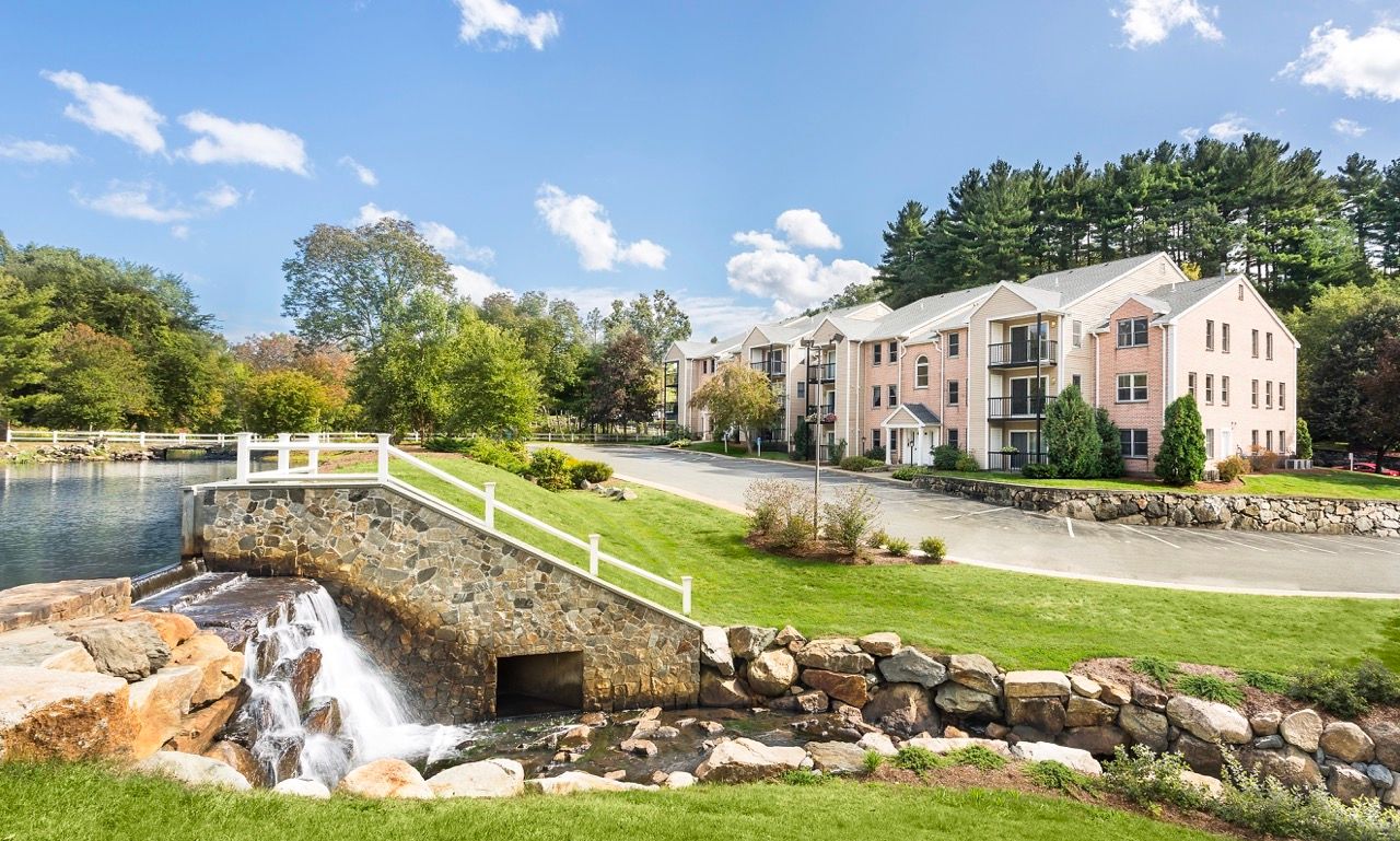 Exterior view of a multi-building apartment community with a stone-lined pond, a small waterfall, and well-kept landscaping.
