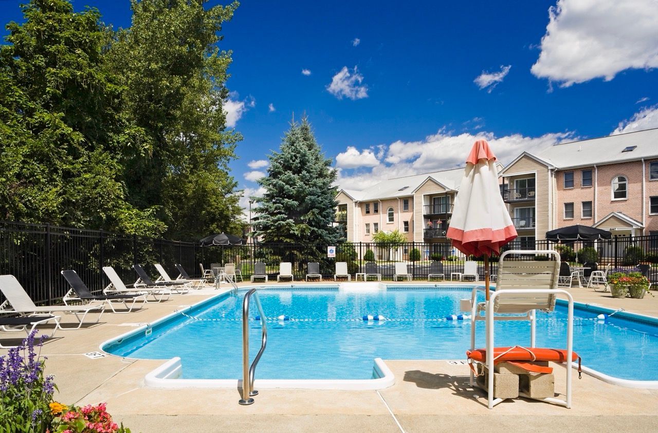 Outdoor community pool area with lounge chairs, umbrellas, and surrounding apartment buildings.