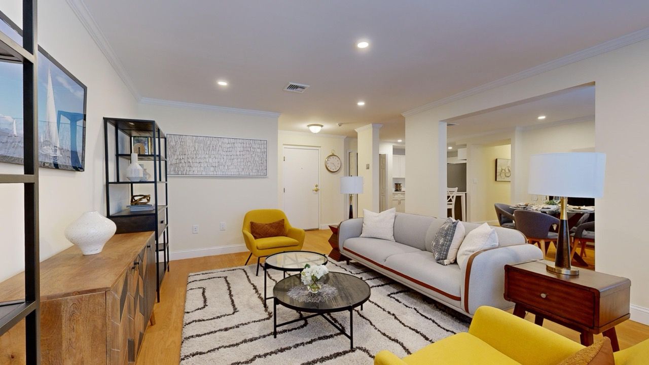 Bright living room with gray sofa, yellow chairs, glass coffee tables, and open dining area.