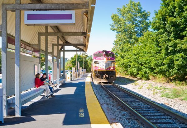 Train approaching a small outdoor station platform with seated passengers and trees beside the tracks