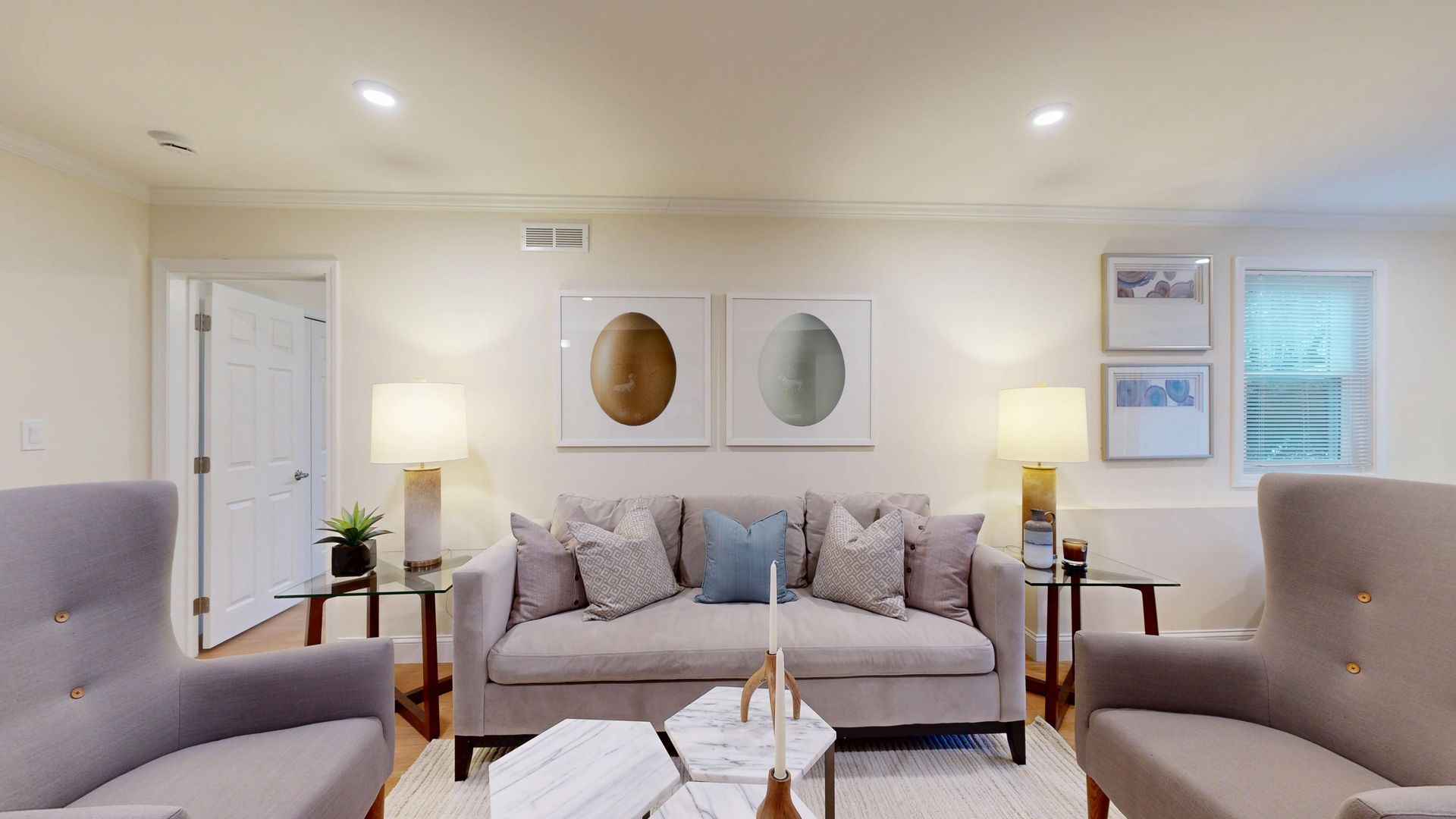Bright living room with beige sofa, armchairs, floor lamps, wall art, and a coffee table in front of a fireplace