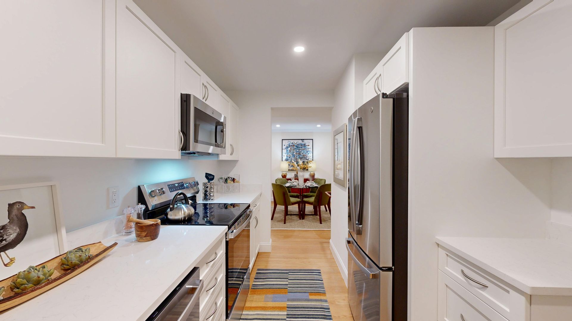 Bright galley kitchen with white cabinets, stainless appliances, striped runner, and dining area in the distance
