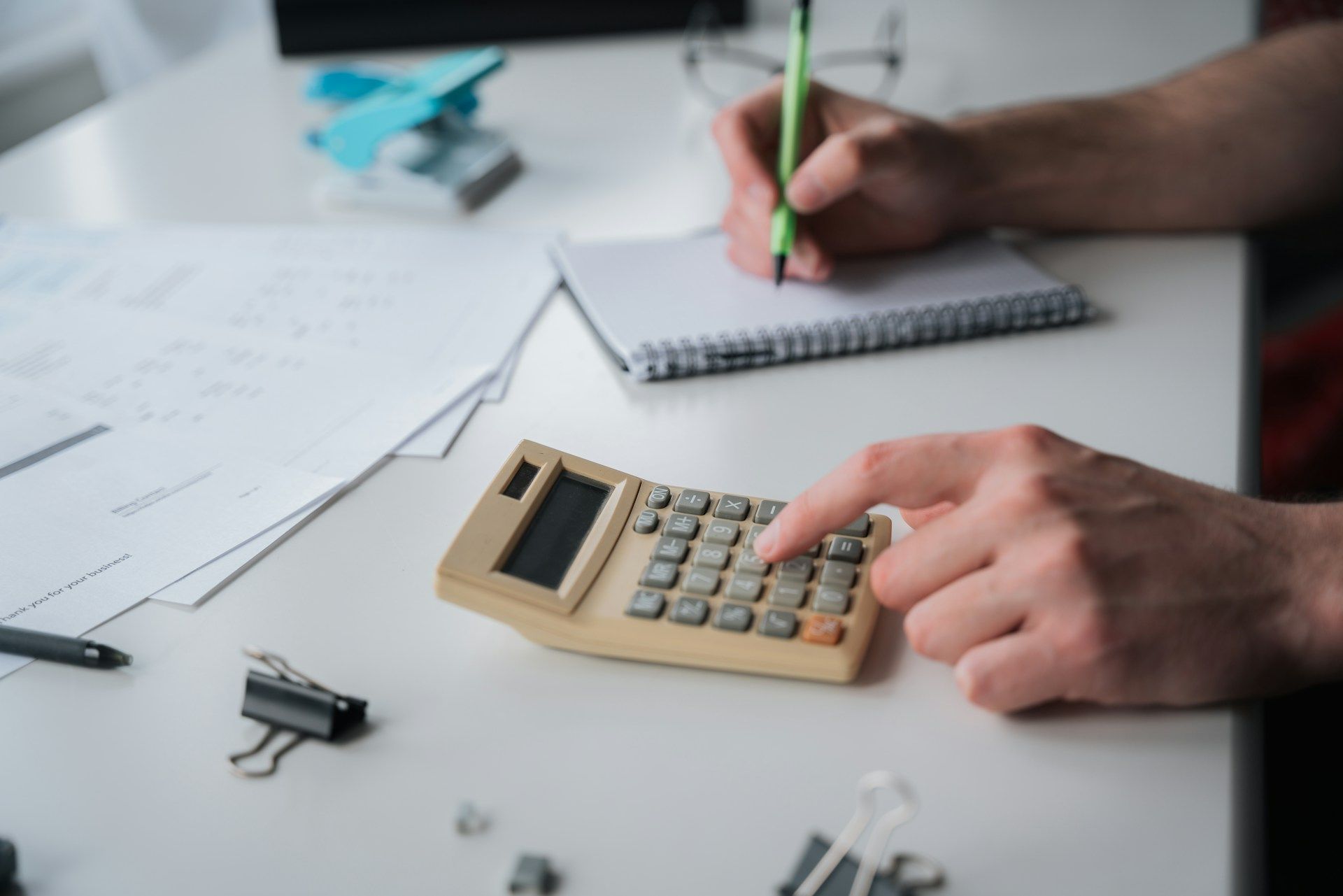 Hands using a calculator and writing in a notebook on a desk with papers and office supplies