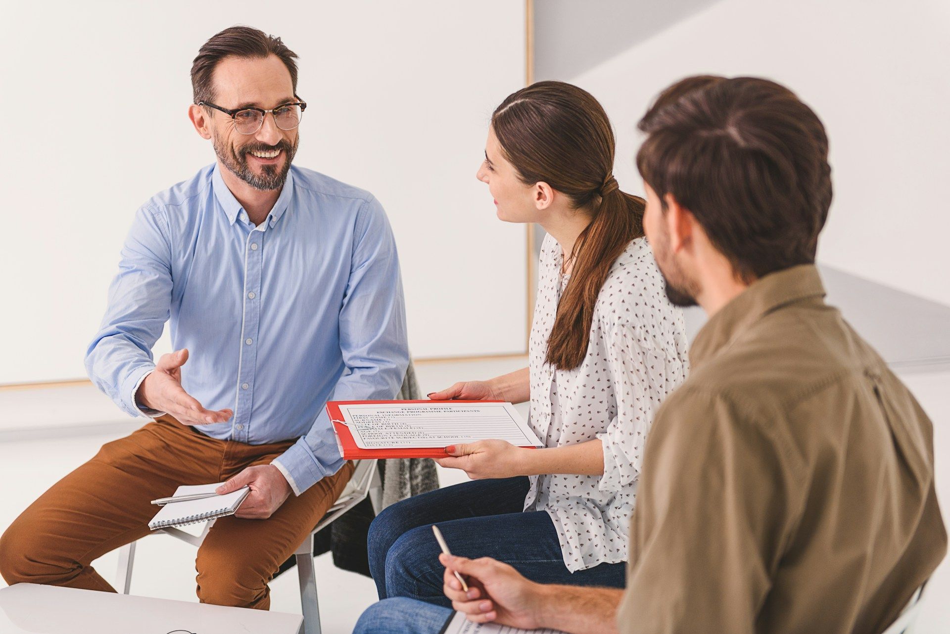 Three people in a meeting, one speaking while others take notes with a clipboard.