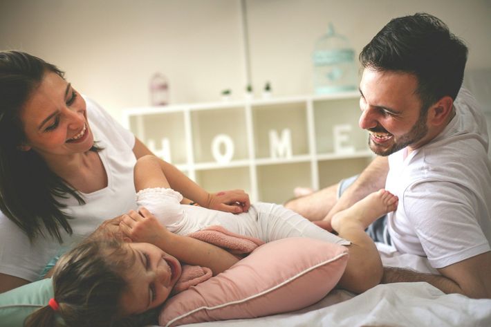 Family smiling on a bed, with a young child between two adults in a bright bedroom