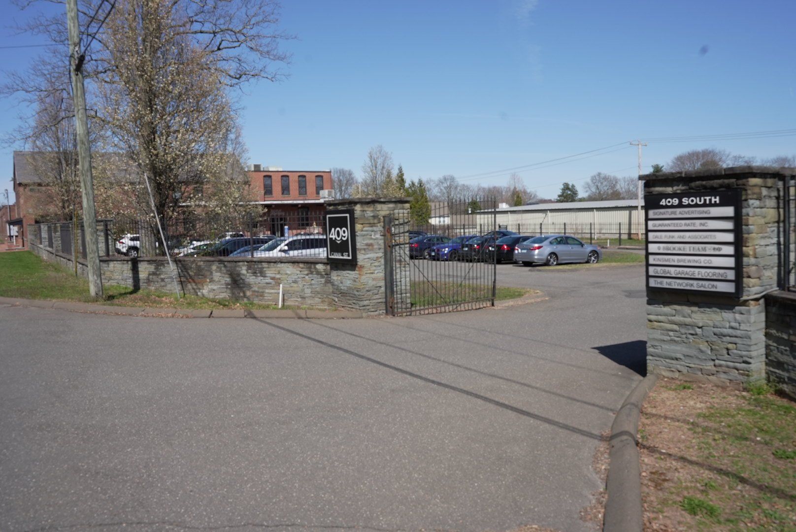 Entrance to a parking lot, marked by stone pillars and a sign. Cars are parked behind a fence.