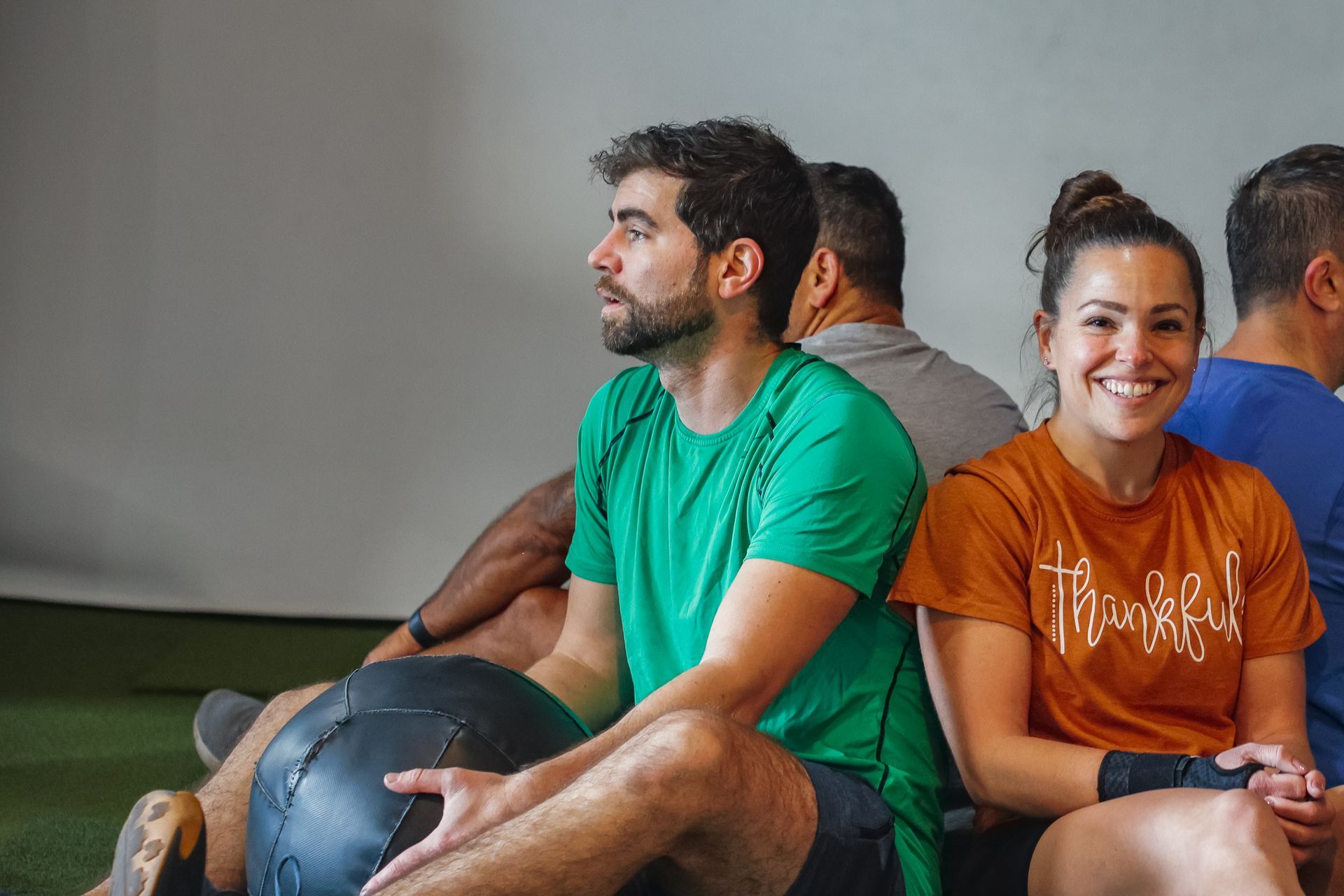 Group of people resting during a workout, one holding a medicine ball, smiling woman, green and orange shirts.