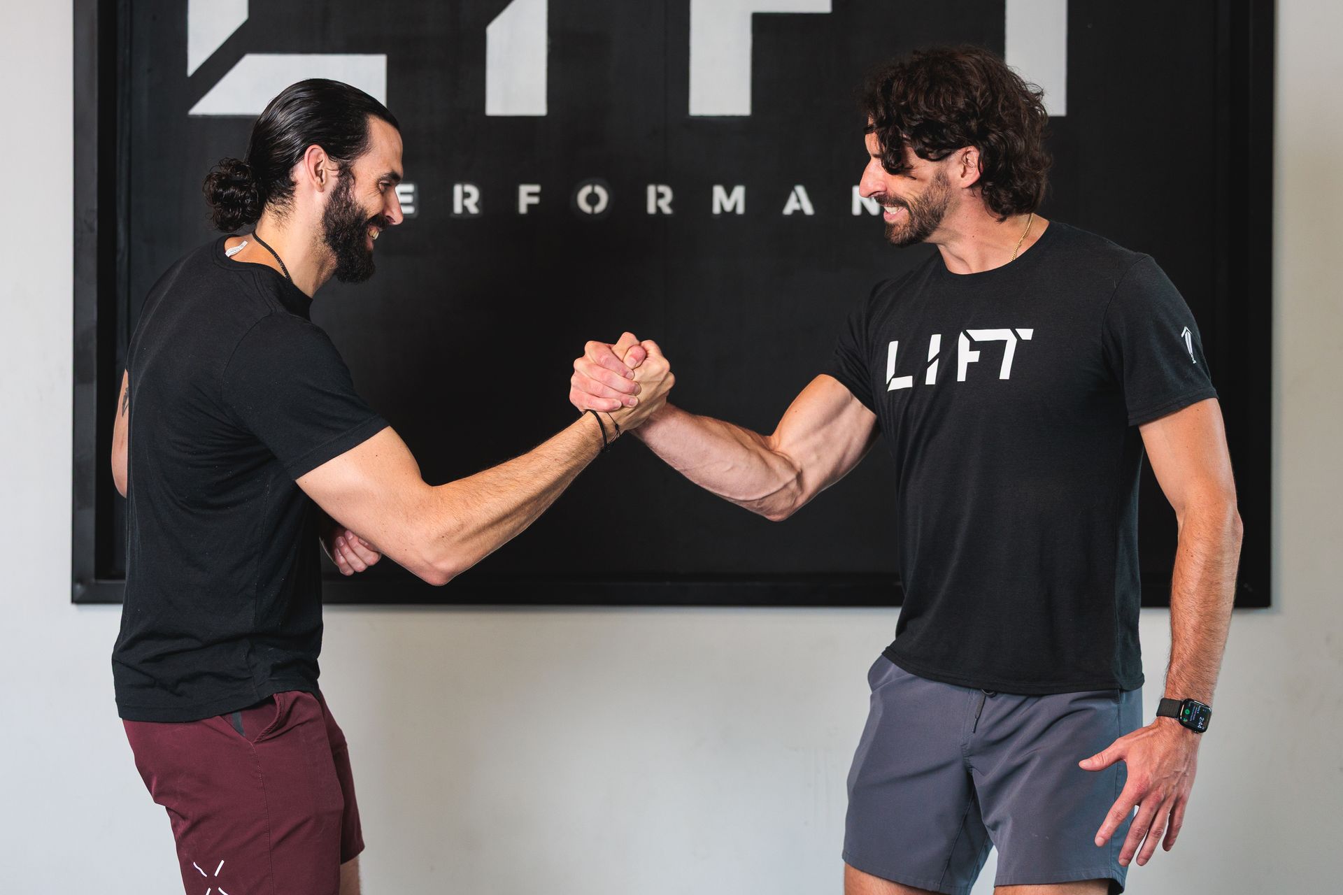 Two men in black shirts arm wrestle in a gym, standing before a dark performance sign.