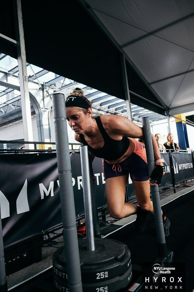 Athlete leaning on a sled in a HYROX competition lane under a tent