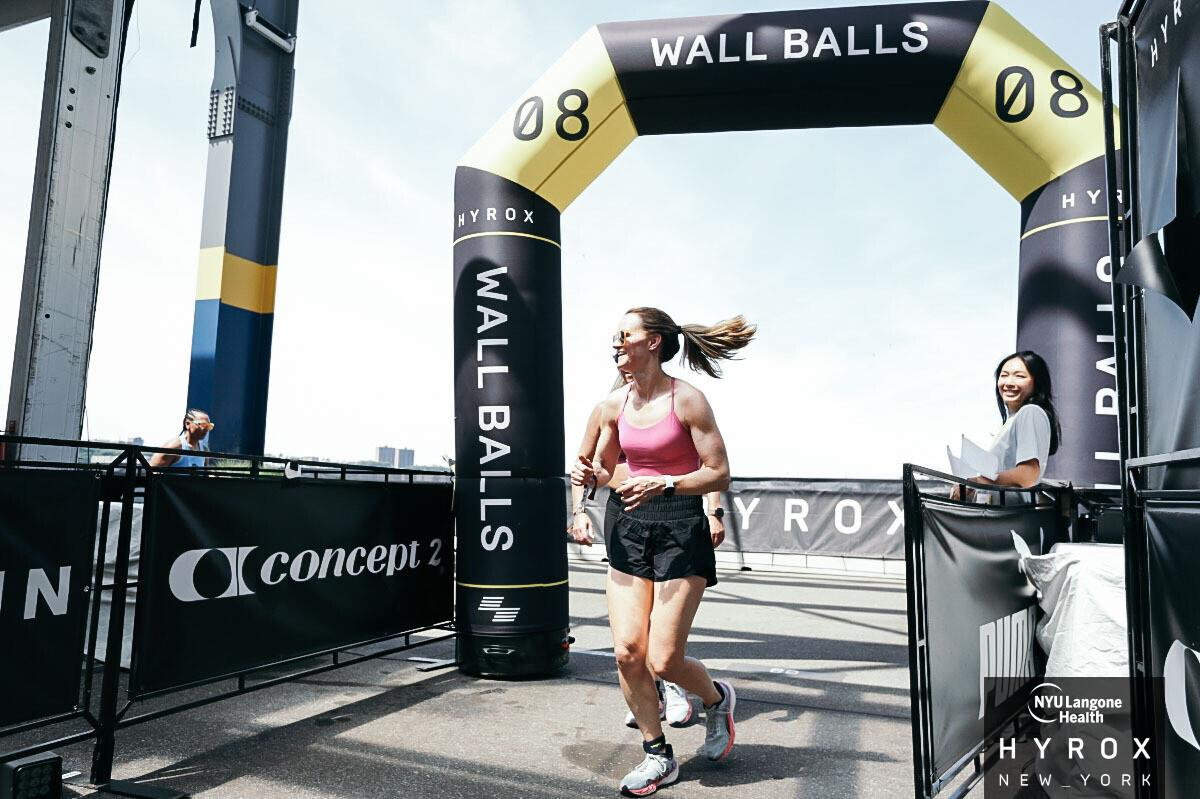 Runner crossing a WALL BALLS finish arch in bright sunlight, with another person nearby