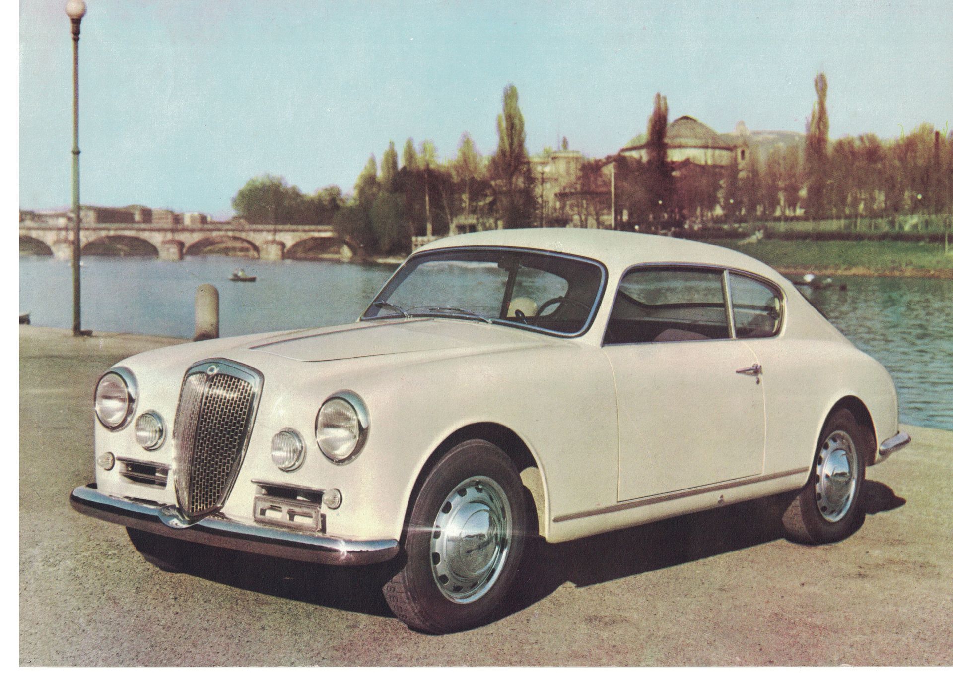 White Lancia Aurelia coupe parked near water with a bridge and building background.