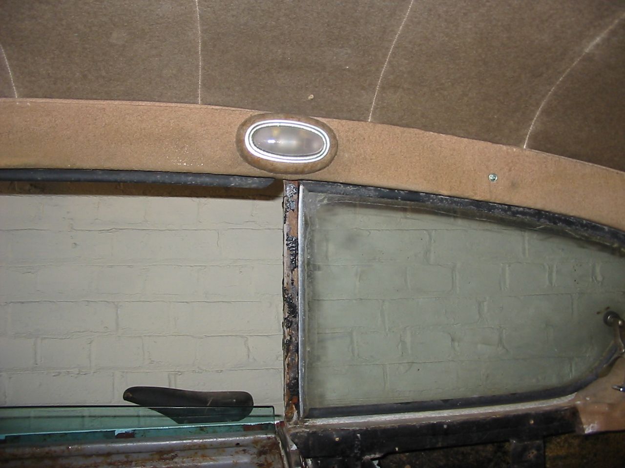 Interior view of a vintage car's window, revealing a light fixture, tan fabric, and a partially exposed brick wall.