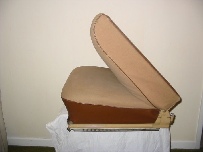 Tan and brown car seat with raised back section, resting on a beige surface, against a light wall.