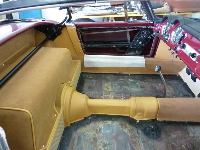 Interior of a vintage car with tan carpet and a painted drivetrain tunnel; door panel removed.