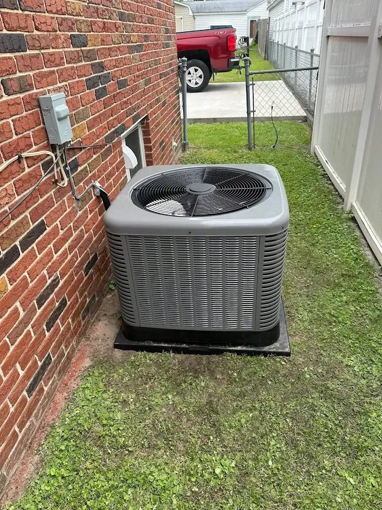 Outdoor air conditioner unit beside a brick house and white fence on a grassy side yard