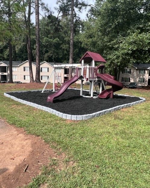 Children's playground with a maroon slide and play structure on a mulch bed in a grassy apartment courtyard