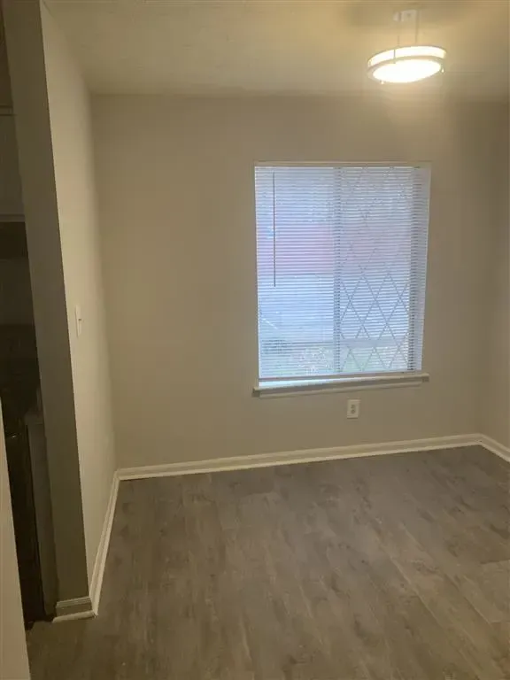 Empty beige room with wood-look flooring, a window with blinds, and a ceiling light fixture.