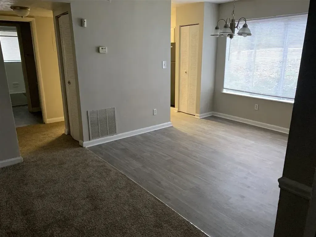 Empty living room with gray carpet, light wood flooring, and large window with blinds
