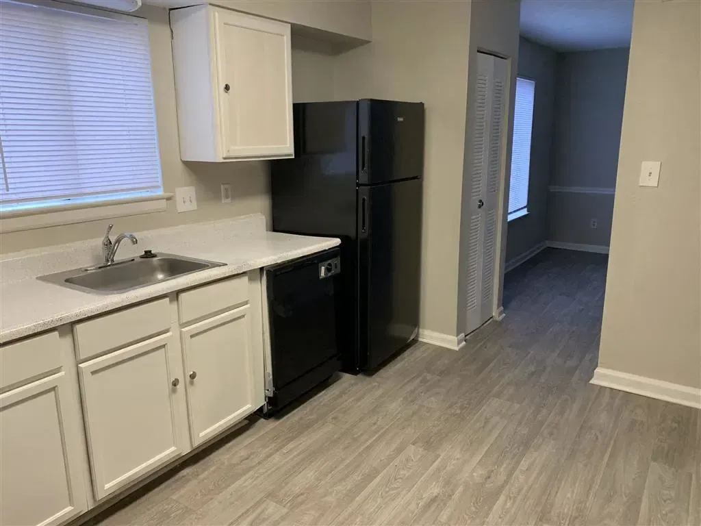 Small kitchen with white cabinets, black refrigerator, and gray wood-look flooring leading to a hallway